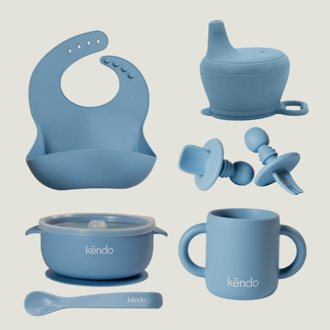 Collection of blue silicone baby products including a bib, cup, bowl, and spoon on a light gray background.